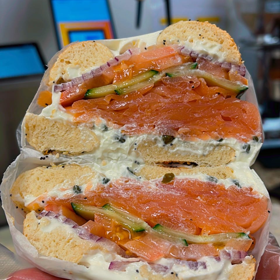 Two sesame bagel sandwiches layered with smoked salmon, cream cheese, cucumber, tomato, and red onion in a restaurant kitchen.