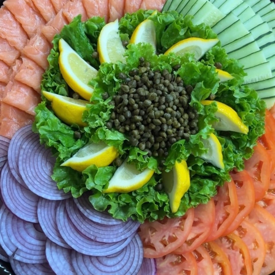 Platter of sliced salmon, cucumber, tomato, red onion, lettuce, lemon wedges, and capers arranged around the center.