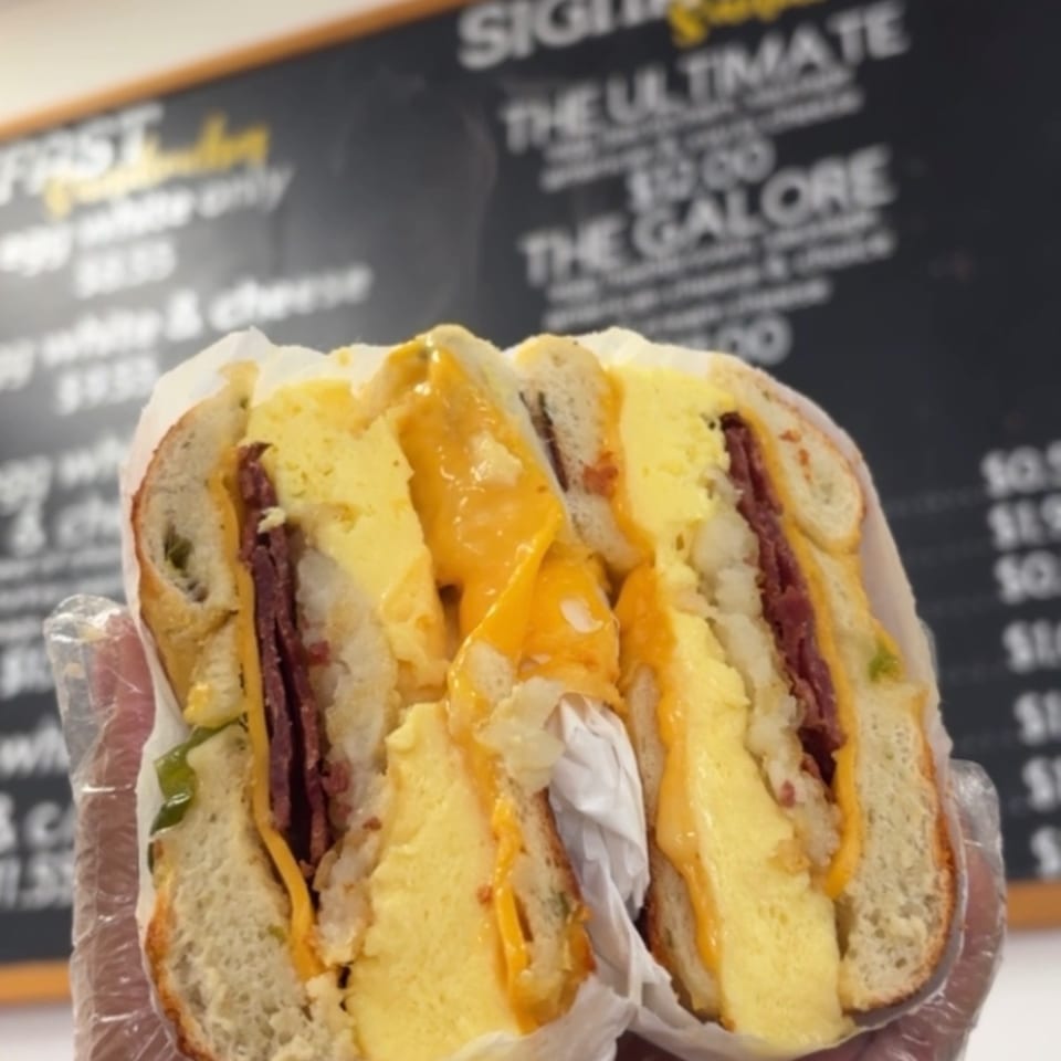 Split bagel sandwich layered with egg, melted cheese, deli meat, and vegetables, held in front of a Bagels Galore menu.