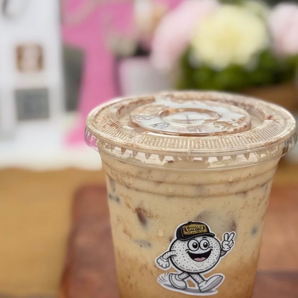 A creamy iced coffee in a clear branded cup sits on a wooden table, with blurred pastel flowers and a menu in the background.