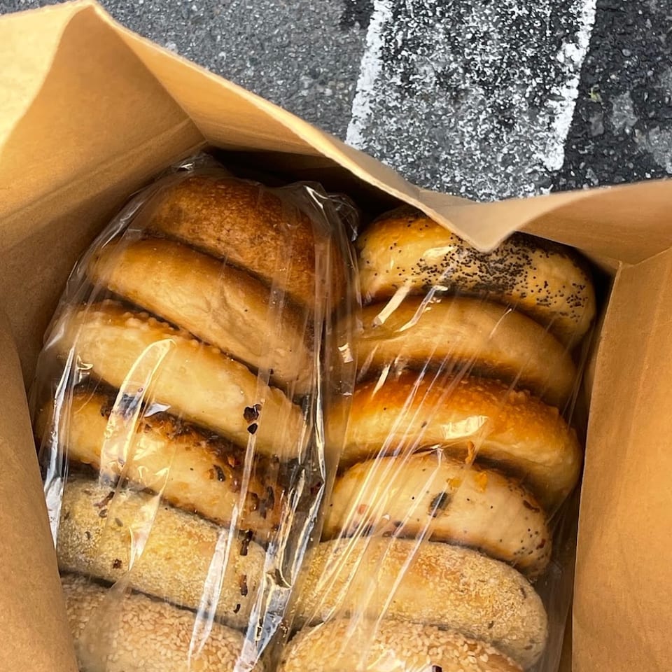 Assorted golden-brown sesame and seeded bagels wrapped in plastic inside a cardboard box on pavement.