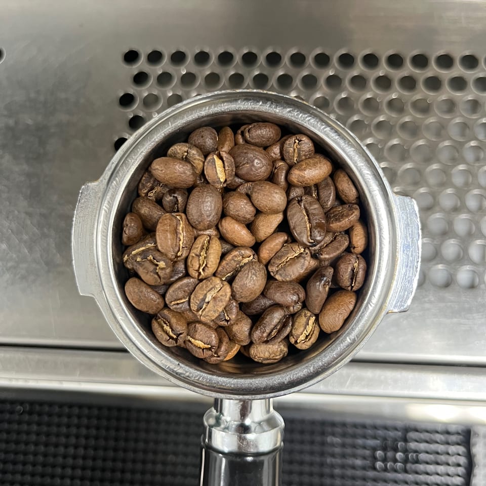 Roasted coffee beans fill the hopper of a commercial espresso grinder on a stainless-steel counter.