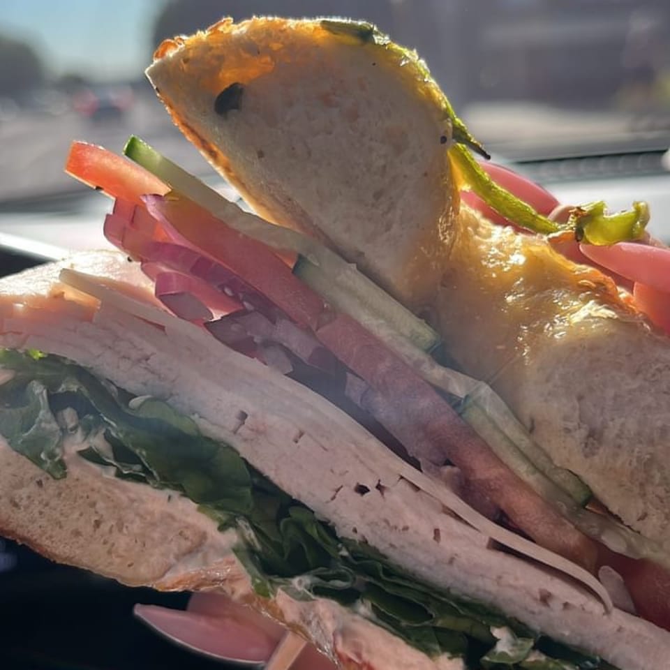 Close-up of a deli sandwich with turkey, lettuce, tomato, cucumber, and red onion, held inside a car.